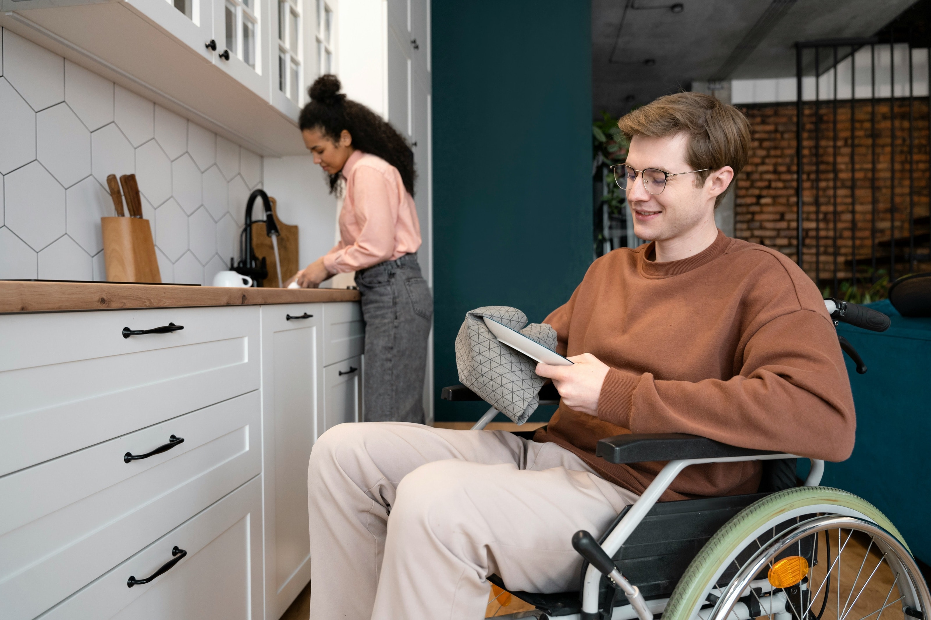 A young man in a wheelchair reads his tablet in a kitchen while a friend washes up behind him.